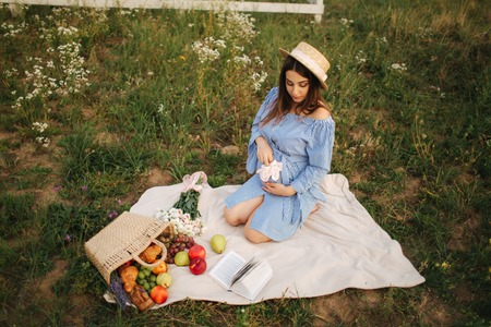 Charming pregnant woman sits in te fieln on the plaid. Woman dressed in blue dress and knitted hat. She take a fruit basket. Background of farmの写真素材