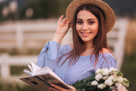 Elegant woman hold a book and bouquet of flower in hand. She stand in the field. Background of farm. Reading the book about babyの写真素材