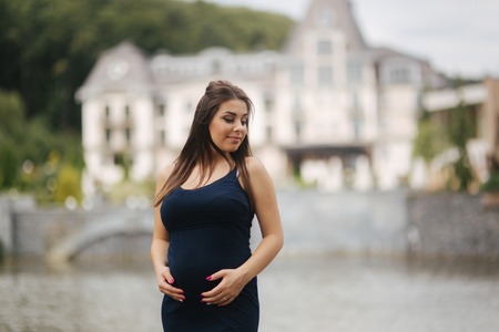 Beautiful young cheerful pregnant woman standing in background of fountain. six months of pregnancy. She pu her hands on her bellyの写真素材
