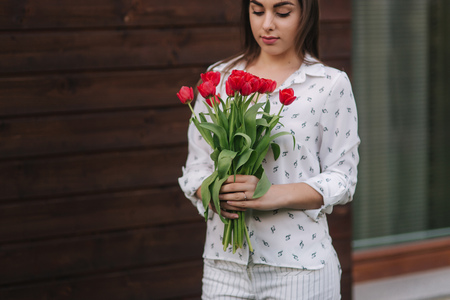 Beautiful girl with bouquet of red flowers stand by the house. Wooden backgroundの写真素材