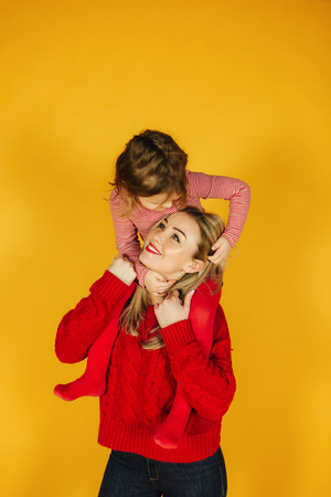 Happy little girl with her young mother on the yellow background. Daughter sitting on moms neck. Smileの写真素材