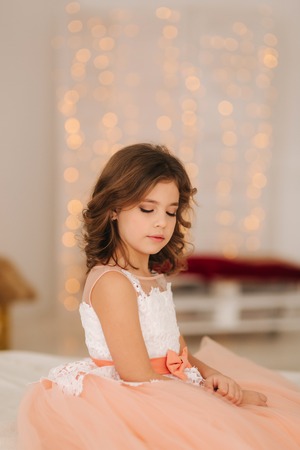 Little beautiful girl with brown hair in a Peach-colored dress. Girl sitting on the sofaの写真素材