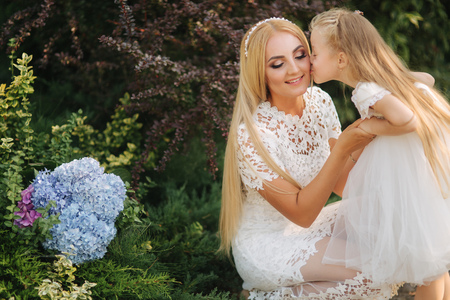 Mom with daughter in white dresses spend time in the park. Blond hair female. Mother weaves pigtails daughter outsideの写真素材