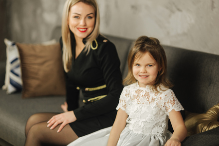 Young mom and little daughter at home sitting on sofa. Attractive mother in black dress. Happy familyの写真素材