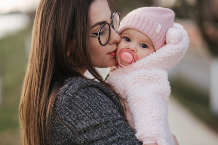 Portrait of mother and her little baby girl. Beautiful mom and cute baby. Mother hud her daughter. Baby dressed in peanch colour hat and nipple toy. Mom kiss her daughterの写真素材