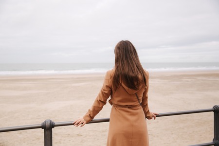 Back view of woman in brown coat standing along North Sea and looking at the wavesの写真素材