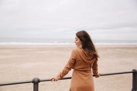 Back view of woman in brown coat standing along North Sea and looking at the wavesの写真素材