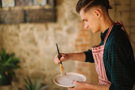 Young artist making ornament on ceramic plate. Handsome young man working at his workshopの写真素材