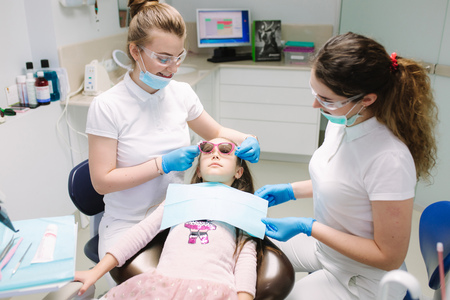 Little girl in 3D glasses sitting on destal chair at the office and have yearly checkup. Dentist with assistantの写真素材
