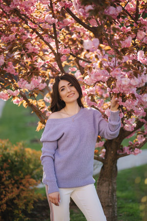 Attractive young woman in a purple sweater stand in front of sakura tree in the park