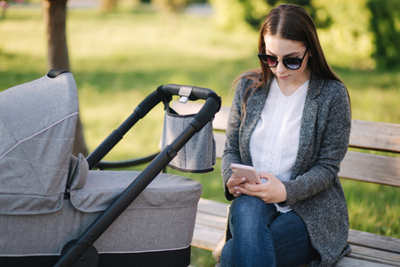 Young mom sitting on the bench with her baby in stroller and use phone. Mothe look something in internetの写真素材
