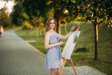 Beautiful girl artist is standing in the park in front of picture and holding palette with paintsの写真素材