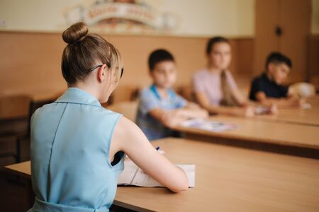Female teacher in front of children. Pretty teacher in classroom sitting at the desk and asking children. education, elementary school, learning and people conceptの写真素材