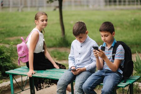 Two boys and girl use their phones during school breack. Cute boys sitting on the bench and play online gamesの写真素材