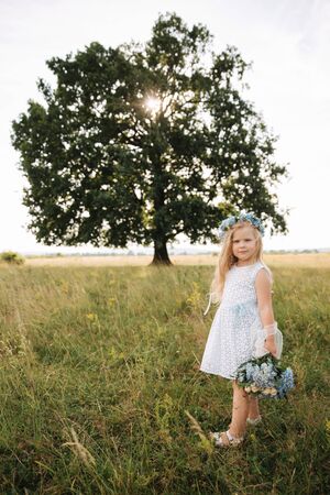 Little girl in sky blue dress with wreath and bouquet stand in field in front of big treeの写真素材