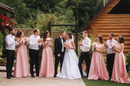 Groom and bride stand with groomsman and bridesmaid outside. Newlyweds kissing and friend clap. Wedding dayの写真素材
