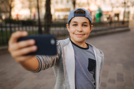 Young boy make a selfie on smartphone in the centre of the city. Cute boy in blue hat. Stylish...