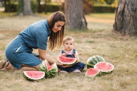 Mom and daughter in demin dresses eat watermelon in summer park. Baby girl sits on grass. Happy family spend time togetherの写真素材