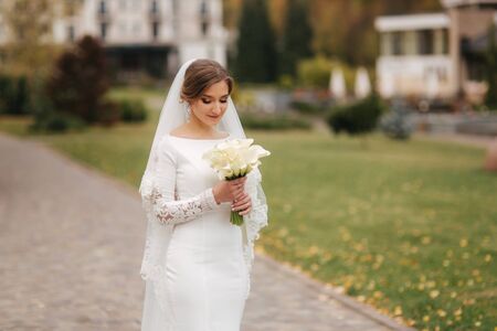 Beautiful bride in elegant wedding dress stand in the park and hold a bouquet of flowers. Portrait of charming ladyの写真素材