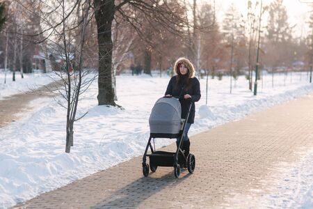 Beautiful mother walking in the park with her little baby in stoller. Woman dressed in blue jaket with hood and jeans. Warm bootsの写真素材
