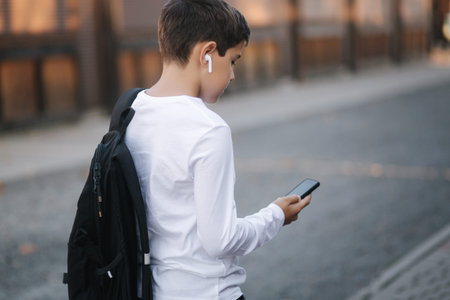 Teenage boy in white sweatshirt and with black backpuck use phone and wireless headphones and print...