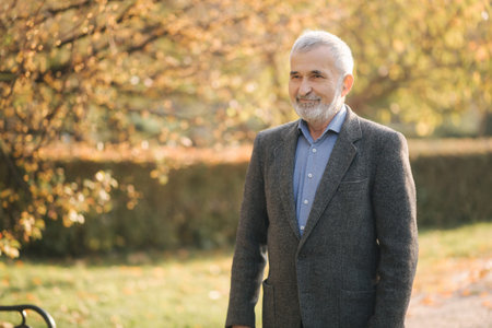 Handsome elderly man putting on a gray jacket. Old gray-haired bearded man walk in the autumn park....