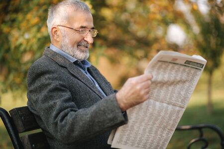 Bearded elderly man in glasses reading newspaper in the autumn park. Handsome gray-haired man sitting on the bench early in the morningの写真素材