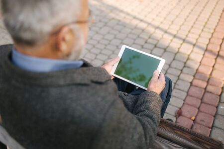Mock up of senior man using tablet outside. Back view of elderly man sitting on the bench and using tabletの写真素材