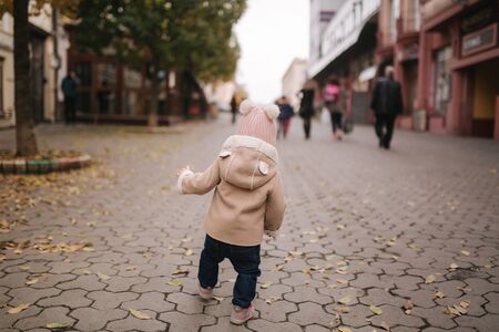Back view of Adorable little baby girl in beautiful brown coat walking in the city. Cute eleven month baby smile and runの写真素材