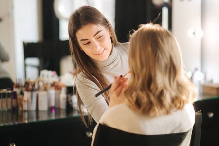 Beautiful female makeup artist doing makeup for a young blond hair girl in a beauty salon sitting in front of a large mirror. Concept of preparation for the holiday and meeting. Start workingの写真素材