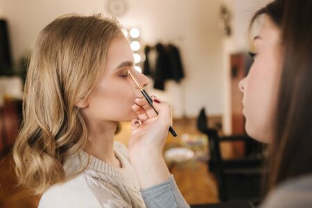 Makeup artist work in her beauty studio. Portrait of Woman applying by professional make up master. Beautiful make up artist start making a makeup for blond hair modelの写真素材
