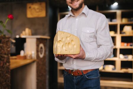 Hansome man hold big slice of cheese maasdam in hand. Cheese with big holes. Background of shelves with cheeseの写真素材