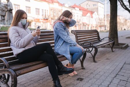 Girl in masks. Coronavirus theme. Women in the city sits on the bench without protective mask, during quarantine. Female sneeze and coughingの写真素材