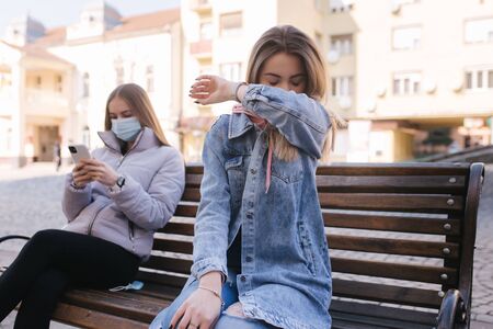 Girl in masks. Coronavirus theme. Women in the city sits on the bench without protective mask, during quarantine. Female sneeze and coughingの写真素材
