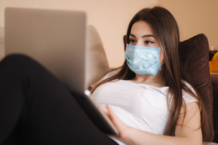 Woman in protective mask working from home during Coronavirus. Female self isolated at home in room....