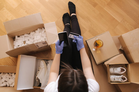 Top view of women in mask and gloves working at home on smartphone on wooden floor with parcel....