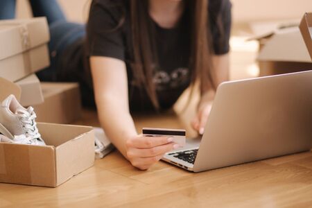 Young woman doing online shopping at home. Female lying on the wooden floor with different parcel. Woman using laptop and credit cardの写真素材