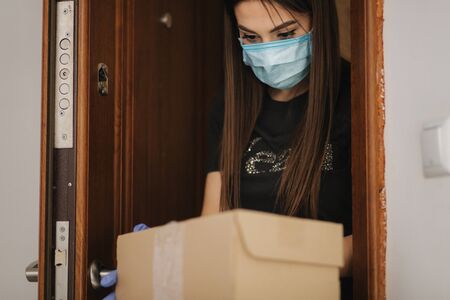 Young woman in maks and gloves take a parcel by her self at home. Package on the floor by the door. Self-qurantine. Coronavirus conceptの写真素材