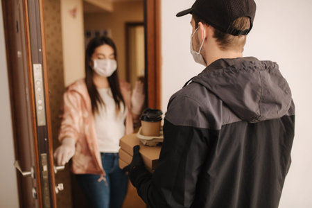 Man delivery pizza and coffe to customer in mask and gloves. Woman open door and take order. Coronavirus themeの写真素材