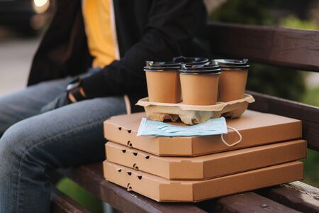Close up Courier in protective mask and medical gloves delivers pizza and coffe. Delivery boy sit on bench and wait for customer. Delivery service under quarantine. Coronavirus covid-19 themeの写真素材