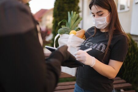 Smart food delivery service. Man in medical mask and gloves handing fresh food to young woman customer receiving express delivery from courier at home. Courier with tablet, sign in. Female order food delivery. Online shopping conceptの写真素材