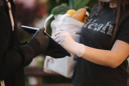 Smart food delivery service. Man in medical mask and gloves handing fresh food to young woman customer receiving express delivery from courier at home. Courier with tablet, sign in. Female order food delivery. Online shopping conceptの写真素材