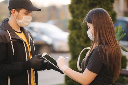 Smart food delivery service. Man in medical mask and gloves handing fresh food to young woman customer receiving express delivery from courier at home. Courier with tablet, sign in. Female order food delivery. Online shopping conceptの写真素材