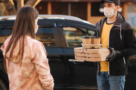 Young woman in protective mask and medical mask take order from delivery boy outside. Food delivery in quarantine. Courier bring Pizza and coffee for customerの写真素材