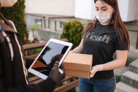 Man from delivery service in medical mask and gloves handing fresh food to young woman customer receiving express delivery from courier at home. Courier with tablet, customer sign in. Female order parcel delivery. Online shopping conceptの写真素材