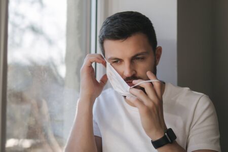 Young man sits on windowsill and out on protective mask and look s out the window. Alone at home in quarantine. Coronavirus theme. Stay at homeの写真素材