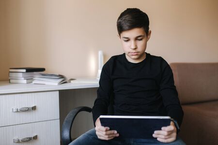Teenage boy at home sits by the table and use tablet during break. Young boy play online games on tablet. Backgroung of desk with booksの写真素材