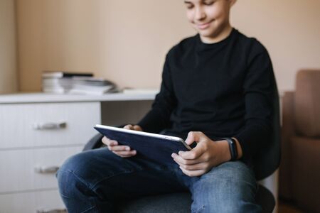 Teenage boy at home sits by the table and use tablet during break. Young boy play online games on tablet. Backgroung of desk with booksの写真素材