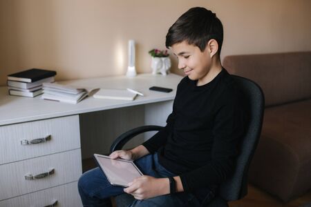 Teenage boy at home sits by the table and use tablet during break. Young boy play online games on tablet. Backgroung of desk with booksの写真素材