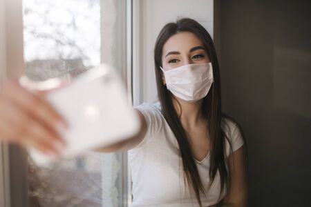 Attractive woman in medical mask make selfie at home. Female in white tshirt and white medical mask use phoneの写真素材